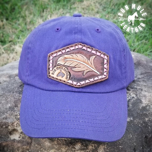 Hand tooled leather patch on soft comfy purple cap. The patch depicts a hand tooled feather with a tooled pink ribbon border. 